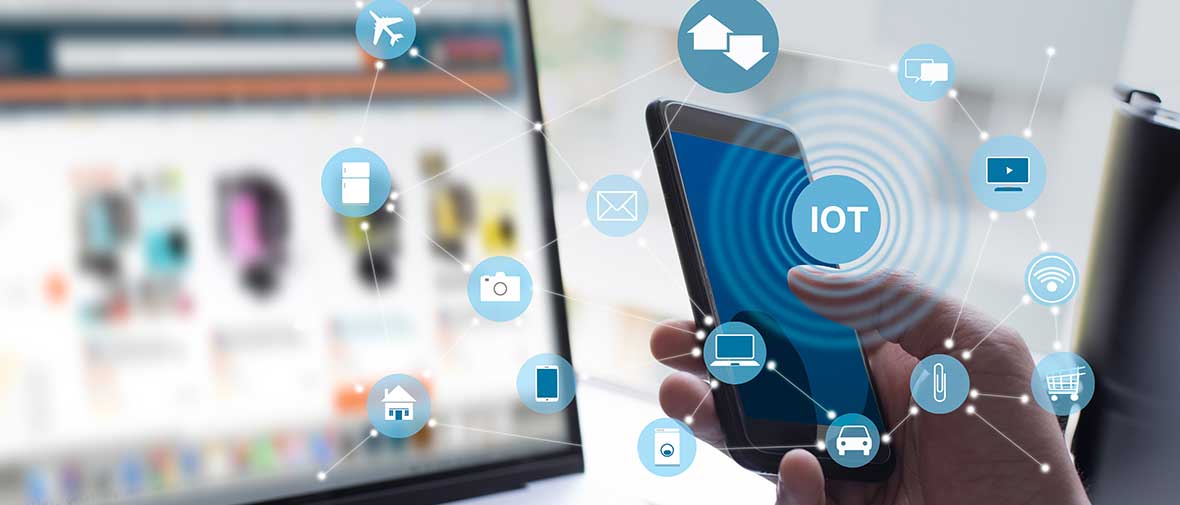 Eine Hand bedient ein Smartphone. Im Hintergrund ein Notebook. Man kann das Wort IoT lesen.